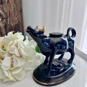 May include: Black ceramic cow creamer with gold accents. The cow is standing on a gold-rimmed oval base.
