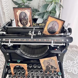 May include: A vintage black typewriter with antique photographs and decorative greenery. The typewriter has a classic design with round keys and a tabulator. The photographs are in ornate frames, adding a touch of nostalgia. The word "Underwood" is visible on the typewriter.