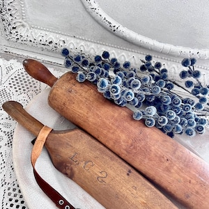 May include: Two vintage wooden rolling pins with dark brown handles and a light brown body. One pin has the initials "L C 2" carved into the wood. A sprig of blue berries rests on top. The rolling pins are on a white decorative plate.