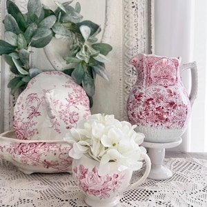 May include: A collection of vintage-style ceramic pieces with a red floral pattern. Includes a pitcher on a pedestal, a small cup with white flowers, and a covered dish. A green leafy wreath is in the background.
