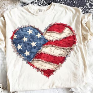 May include: Cream-colored t-shirt featuring a heart-shaped design resembling the American flag. The heart is divided into blue with white stars and red and white stripes, with a distressed, frayed edge.