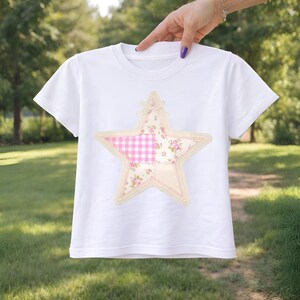 May include: White t-shirt with a star applique. The star is made of pink gingham, floral, and solid fabric pieces, outlined with a cream-colored trim. The shirt has short sleeves and a crew neck.