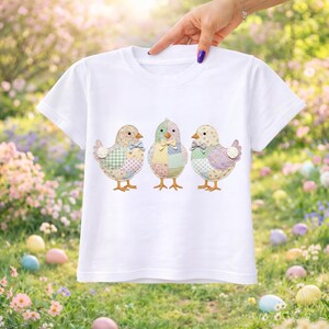 以下が含まれることがあります： 3羽の刺繍されたひよこが特徴の白いTシャツ。それぞれのひよこはパッチワーク風の体と蝶ネクタイをしています。背景はぼやけており、草、花、イースターエッグがあります。Tシャツは手で持たれています。