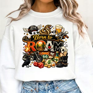 May include: White sweatshirt with a colorful graphic design featuring a motorcycle, snake, beer, dice, and playing cards. The text "Born to Roam" is prominently displayed above the other graphics. The design includes a Route 66 sign.