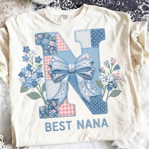 May include: A cream-coloured t-shirt with a large, appliquéd letter "N" in blue and pink floral patterns and a bow. The text "BEST NANA" is below the letter. Floral accents are on either side of the letter, creating a decorative design.