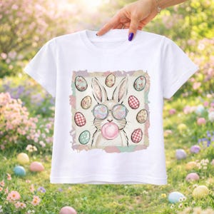 Puede incluir: Camiseta blanca con un conejo de dibujos animados con gafas de sol y soplando una burbuja. El conejo está rodeado de huevos de Pascua con estampados florales y a cuadros. La camiseta está sujeta por una mano.