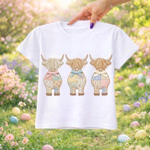May include: White t-shirt with a design of three Highland cows. Each cow has a patchwork body and a different colored bow. The background is a blur of flowers and Easter eggs, creating a spring theme.