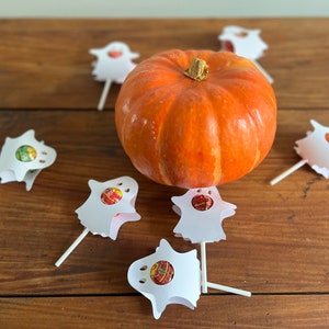 Puede incluir: Una peque&ntilde;a calabaza naranja se encuentra sobre una mesa de madera rodeada de seis piruletas blancas con forma de fantasma con centros de caramelo de colores.
