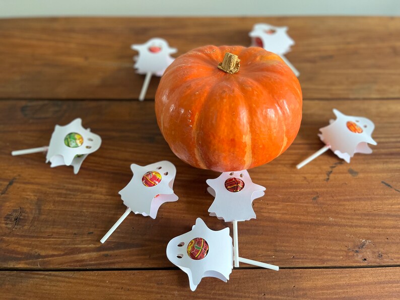 Puede incluir: Una peque&ntilde;a calabaza naranja se encuentra sobre una mesa de madera rodeada de seis piruletas blancas con forma de fantasma con centros de caramelo de colores.