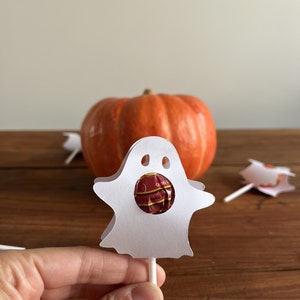 Puede incluir: Lollipop de papel blanco en forma de fantasma con un lollipop rojo y blanco en su interior. El fantasma est&aacute; sobre una superficie de madera con una calabaza en el fondo.