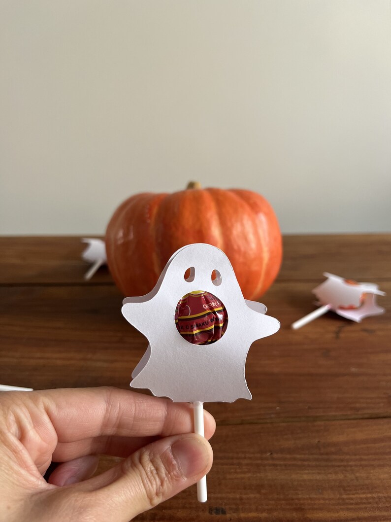 Puede incluir: Lollipop de papel blanco en forma de fantasma con un lollipop rojo y blanco en su interior. El fantasma est&aacute; sobre una superficie de madera con una calabaza en el fondo.