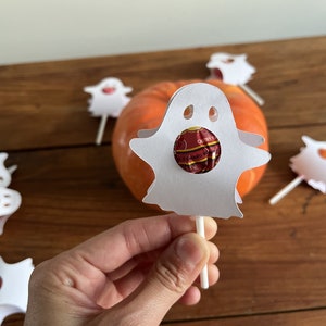 Puede incluir: Piruleta de papel blanco en forma de fantasma con una piruleta roja y naranja en su interior. El fantasma est&aacute; en un palo y hay otros fantasmas en el fondo.