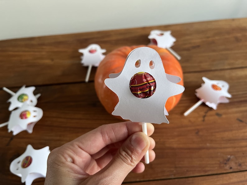 Puede incluir: Piruleta de papel blanco en forma de fantasma con una piruleta roja y naranja en su interior. El fantasma est&aacute; en un palo y hay otros fantasmas en el fondo.