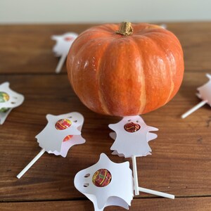 Puede incluir: Una peque&ntilde;a calabaza naranja se encuentra sobre una mesa de madera rodeada de cinco soportes de piruletas con forma de fantasma blancos. Cada soporte contiene un caramelo redondo.