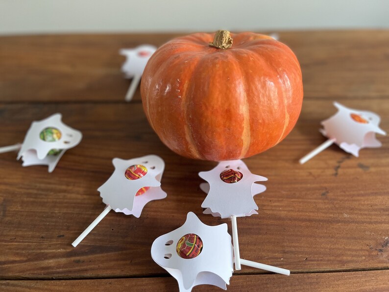 Puede incluir: Una peque&ntilde;a calabaza naranja se encuentra sobre una mesa de madera rodeada de cinco soportes de piruletas con forma de fantasma blancos. Cada soporte contiene un caramelo redondo.