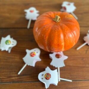 Puede incluir: Una peque&ntilde;a calabaza naranja se encuentra en el centro de una mesa de madera rodeada de siete piruletas fantasma blancas.