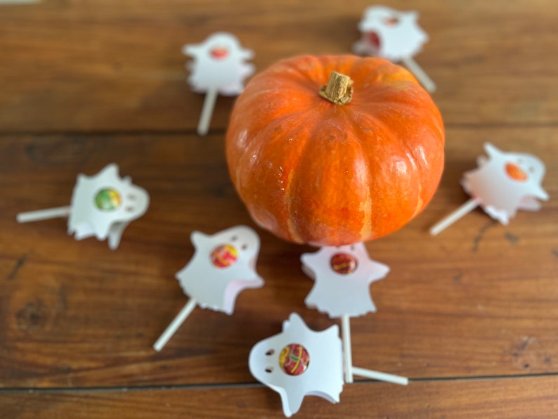 Puede incluir: Una peque&ntilde;a calabaza naranja se encuentra en el centro de una mesa de madera rodeada de siete piruletas fantasma blancas.