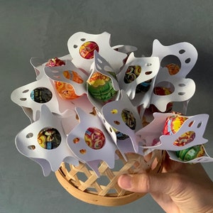 Puede incluir: Porta golosinas de papel blanco en forma de fantasma llenos de piruletas de colores. Los soportes est&aacute;n dispuestos en una cesta.