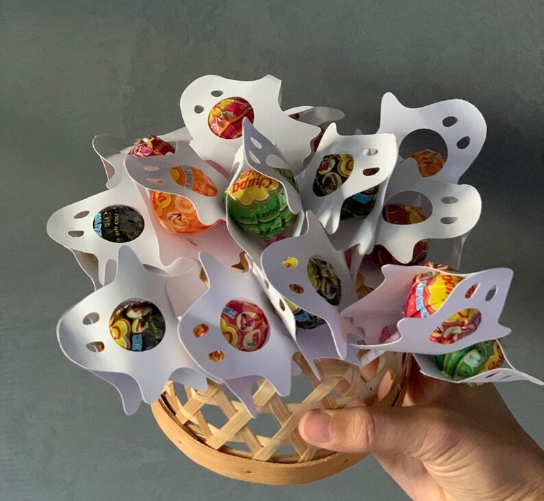 Puede incluir: Porta golosinas de papel blanco en forma de fantasma llenos de piruletas de colores. Los soportes est&aacute;n dispuestos en una cesta.