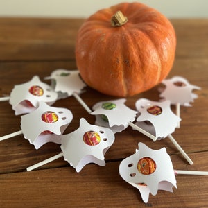Puede incluir: Un grupo de soportes de piruletas con forma de fantasma blanco con piruletas de colores en su interior, sentados sobre una superficie de madera junto a una peque&ntilde;a calabaza naranja.