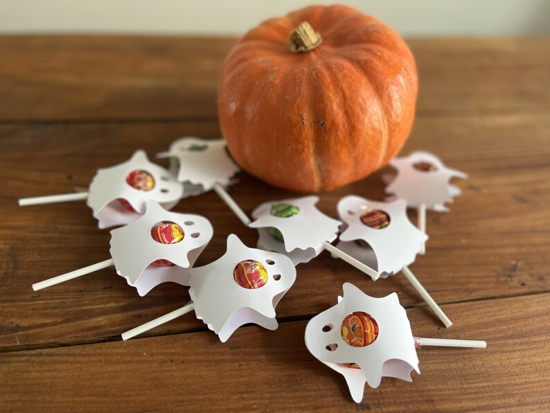 Puede incluir: Un grupo de soportes de piruletas con forma de fantasma blanco con piruletas de colores en su interior, sentados sobre una superficie de madera junto a una peque&ntilde;a calabaza naranja.