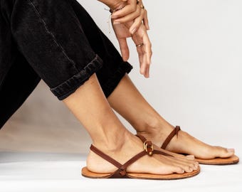 May include: A pair of brown leather thong sandals featuring thin straps, a small gold-toned metal buckle detail at the center, and a flat sole. The sandals are worn with black denim pants, showcasing a minimalist design with simple construction.