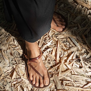 Pode incluir: Um par de sand&aacute;lias de couro castanho escuro com um design minimalista de tiras tipo tanga que envolvem o tornozelo e passam entre os dedos, combinadas com uma pe&ccedil;a de roupa preta comprida que chega at&eacute; aos tornozelos.