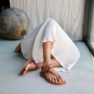 Pode incluir: Um par de sand&aacute;lias de couro castanho com tiras finas que envolvem o p&eacute; e o tornozelo, combinadas com um vestido branco comprido e fluido feito de um tecido texturizado e leve que cai suavemente sobre as pernas e os p&eacute;s.