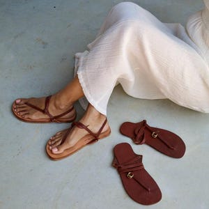 Pode incluir: Um par de sand&aacute;lias de dedo em couro castanho com tiras finas e um pequeno detalhe de anel em tom dourado ao centro. As sand&aacute;lias apresentam uma sola plana e um design de tira envolvente no tornozelo. Duas sand&aacute;lias a condizer est&atilde;o pr&oacute;ximas.