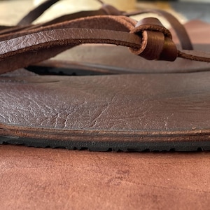 May include: Close-up of a brown leather sandal with a simple design and a black rubber sole.