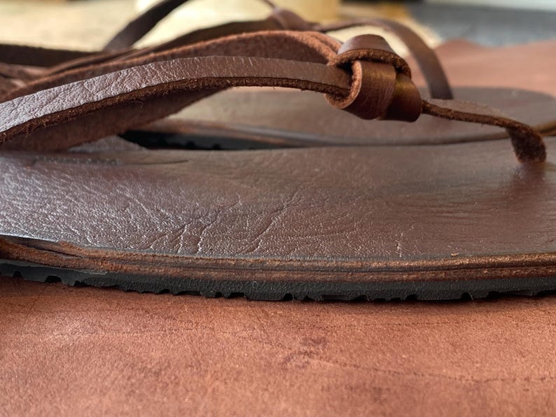 May include: Close-up of a brown leather sandal with a simple design and a black rubber sole.