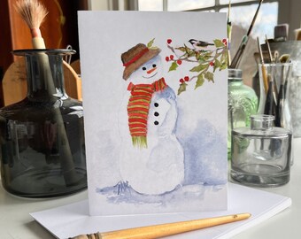 May include: A watercolour painting of a snowman wearing a red and green striped scarf and a brown hat. The snowman is standing in a snowy landscape with a small bird perched on a branch with red berries.