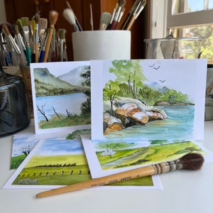 May include: Four watercolour paintings on white cardstock, featuring landscapes with trees, water, and rocks. The paintings are arranged in a stack, with a paintbrush resting on top.
