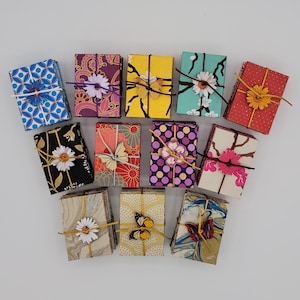 May include: A collection of twelve small rectangular notebooks with various patterned covers, including floral, geometric, and marbled designs. Each notebook is tied with a thin cord and decorated with a small wooden butterfly or flower.
