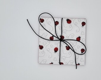 May include: A square gift wrapped in white paper with a ladybug pattern and a black ribbon tied in a bow. The ladybugs are red with black spots. The background is plain white.