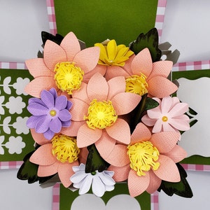 May include: A pink and green explosion box card with a bouquet of paper flowers. The flowers are pink, yellow, white, and purple. The box is decorated with a pink and white gingham pattern.