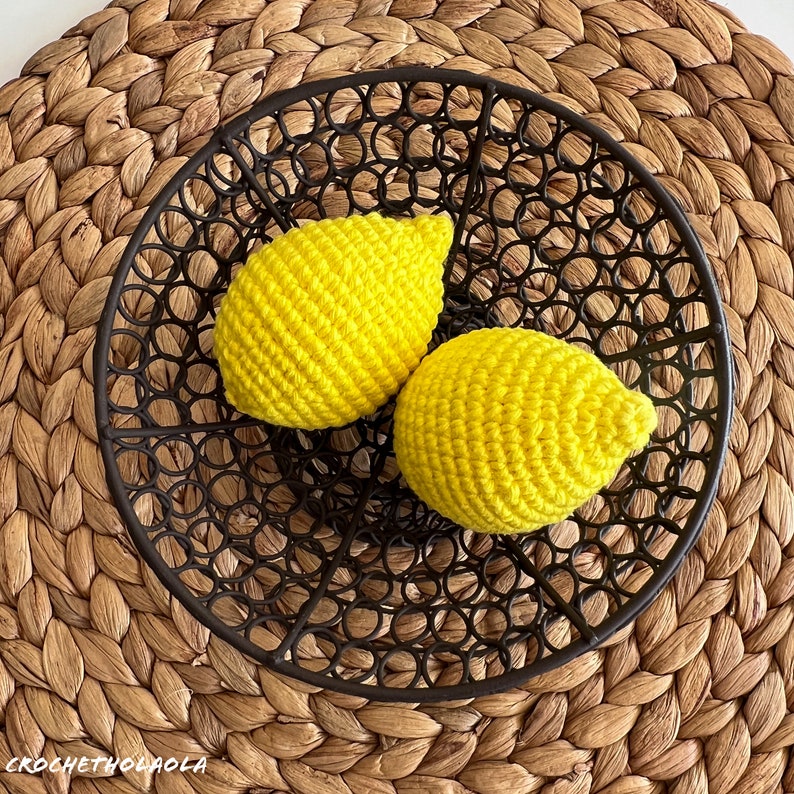 May include: Two crocheted yellow lemons in a black metal bowl. The bowl has a circular design with a woven pattern.