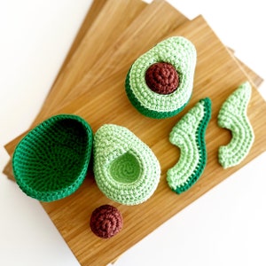 Puede incluir: Un conjunto de juguetes de aguacate de crochet, que incluye un aguacate entero, un hueso y rodajas de aguacate. Los juguetes son verdes y marrones y se muestran en una tabla de cortar de madera.