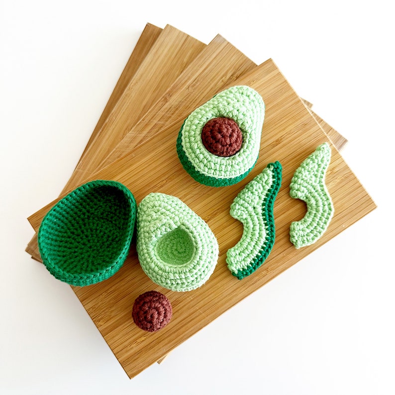 May include: A set of crocheted avocado halves, a whole avocado, and a pit on a wooden chopping board. The avocado halves are green and light green, while the pit is brown.