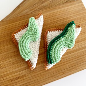 May include: Two crocheted avocado toast ornaments, one with a light green avocado and the other with a darker green avocado, both with brown edges.