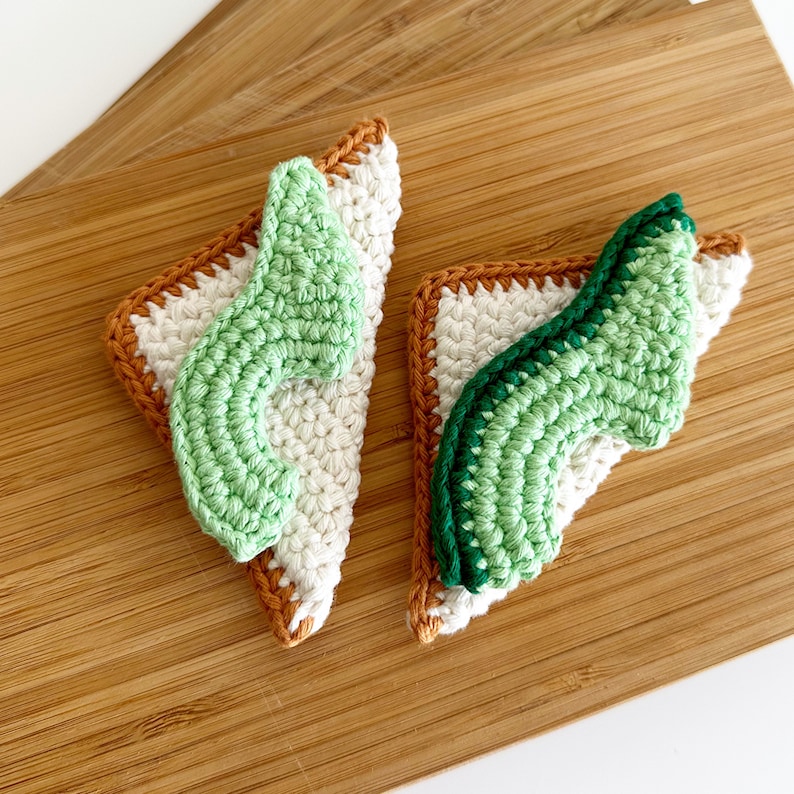 May include: Two crocheted avocado toast ornaments, one with a light green avocado and the other with a darker green avocado, both with brown edges.