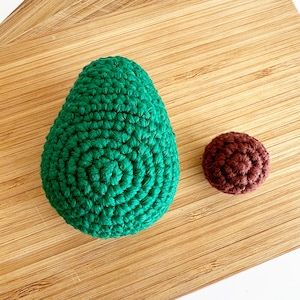 May include: A green crocheted avocado and a brown crocheted pit on a wooden surface.