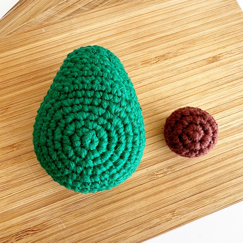 May include: A green crocheted avocado and a brown crocheted pit on a wooden surface.