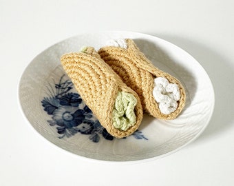 May include: Two crocheted cannoli, one with green filling and one with white filling, on a white plate with blue floral design.