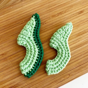 May include: Two crocheted avocado halves in shades of green, one with a dark green border, on a wooden surface.