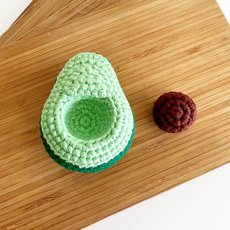 May include: A crocheted avocado with a green and brown colour scheme. The avocado is split open to reveal a brown crocheted pit.