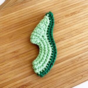 May include: A crocheted avocado half, with a green and dark green colour scheme. The avocado is on a wooden surface.