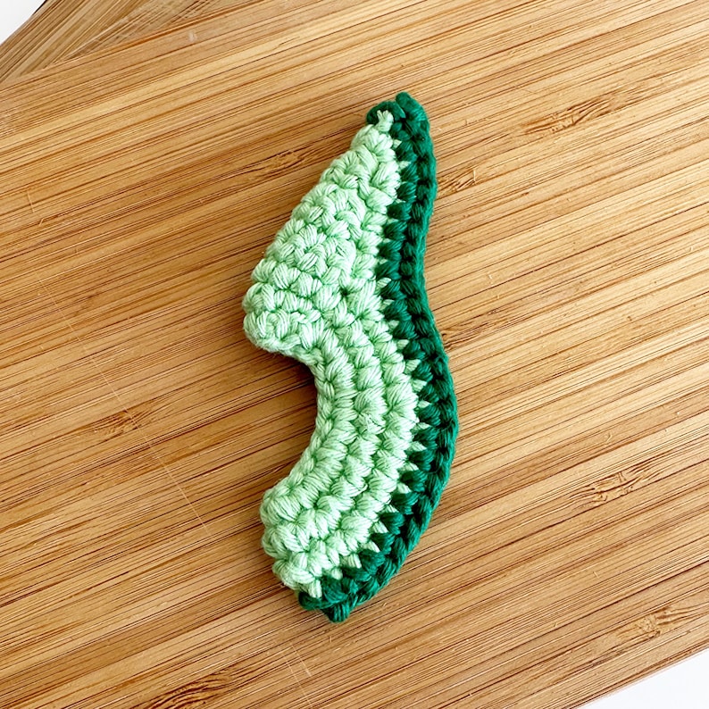 May include: A crocheted avocado half, with a green and dark green colour scheme. The avocado is on a wooden surface.