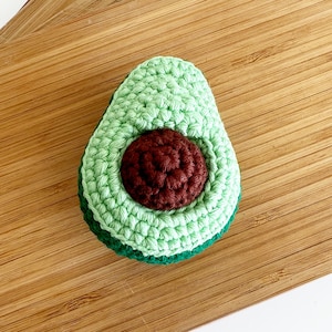 May include: A crocheted avocado toy with a green outer layer and a brown pit. The avocado is sitting on a wooden surface.