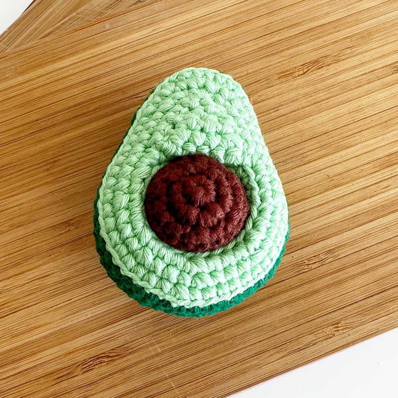 May include: A crocheted avocado toy with a green outer layer and a brown pit. The avocado is sitting on a wooden surface.